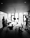 Ein Schwarz-Weiß-Bild zeigt zwei weiße Männer, die sich vor einem Fenster unterhalten, das viel Licht hereinlässt. Der Boden und die Decke des Raums sind dunkel, und in der Mitte des Raums stehen ein schwarzer Tisch mit verschiedenen schwarzen Stühlen und hängt eine dreiköpfige Hängelampe. Ein Schwarz-Weiß-Bild zeigt zwei weiße Männer, die sich vor einem Fenster unterhalten, das viel Licht hereinlässt. Der Boden und die Decke des Raums sind dunkel, und in der Mitte des Raums stehen ein schwarzer Tisch mit verschiedenen schwarzen Stühlen und hängt eine dreiköpfige Hängelampe.