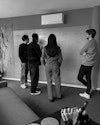 Drei Männer und eine Frau mit langen Haaren stehen vor einem wandgroßen Whiteboard, auf dem Wireframes zu sehen sind, im Vordergrund ein Sofa und Werkstattmaterial. Drei Männer und eine Frau mit langen Haaren stehen vor einem wandgroßen Whiteboard, auf dem Wireframes zu sehen sind, im Vordergrund ein Sofa und Werkstattmaterial.
