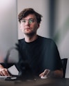 Ein weißer Mann mit schwarzem T-Shirt und runder Brille, dunklem Haar und Dreitagebart sitzt an einem Tisch, schaut nach vorne und bedient mit seiner linken Hand eine weiße Computermaus. Ein weißer Mann mit schwarzem T-Shirt und runder Brille, dunklem Haar und Dreitagebart sitzt an einem Tisch, schaut nach vorne und bedient mit seiner linken Hand eine weiße Computermaus.