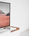 A modern, minimalist entertainment area featuring a game console on a chic wooden shelf, with a large framed landscape screen in front of the white wall above. A modern, minimalist entertainment area featuring a game console on a chic wooden shelf, with a large framed landscape screen in front of the white wall above.